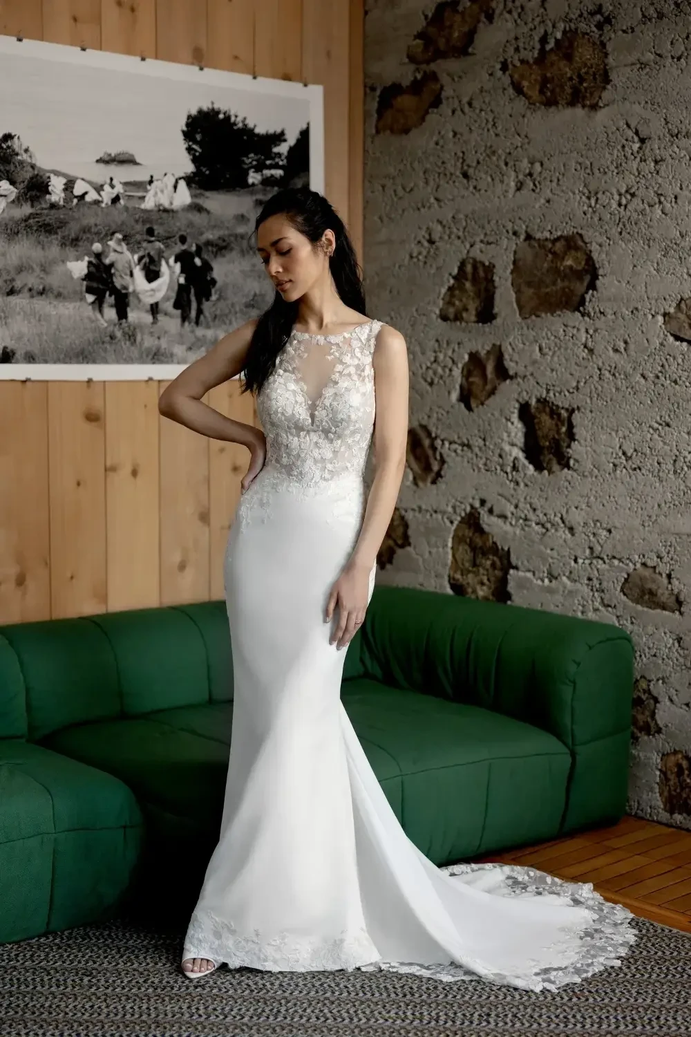 A woman in a fitted white wedding dress stands in a room with wooden walls and a green couch, gazing down thoughtfully. The dress features lace detailing on the bodice and has a train. A black and white photograph is visible in the background.
