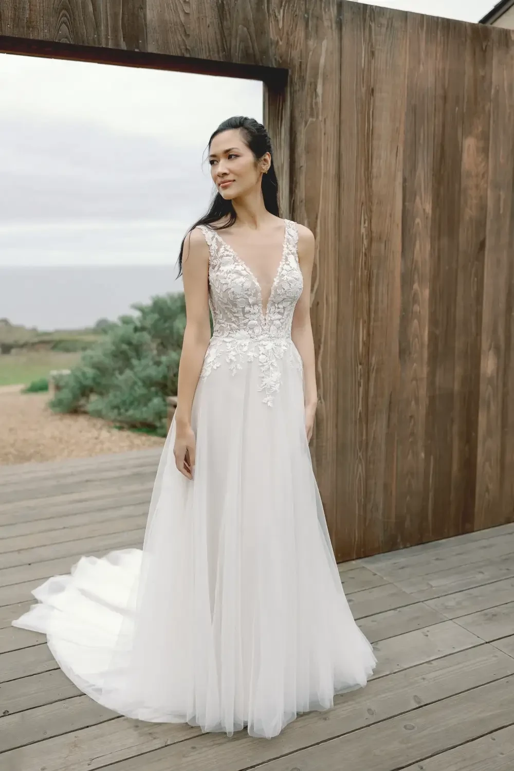 A woman in a white wedding dress stands on a wooden deck, with a natural landscape in the background.