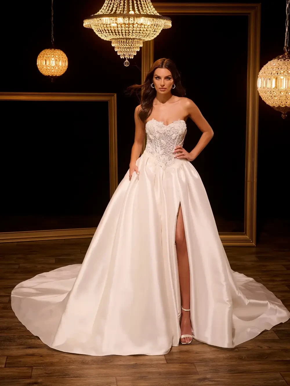 A model in a strapless wedding gown with a glittering bodice and a high slit, standing in a dimly lit room with chandeliers and gold accents.