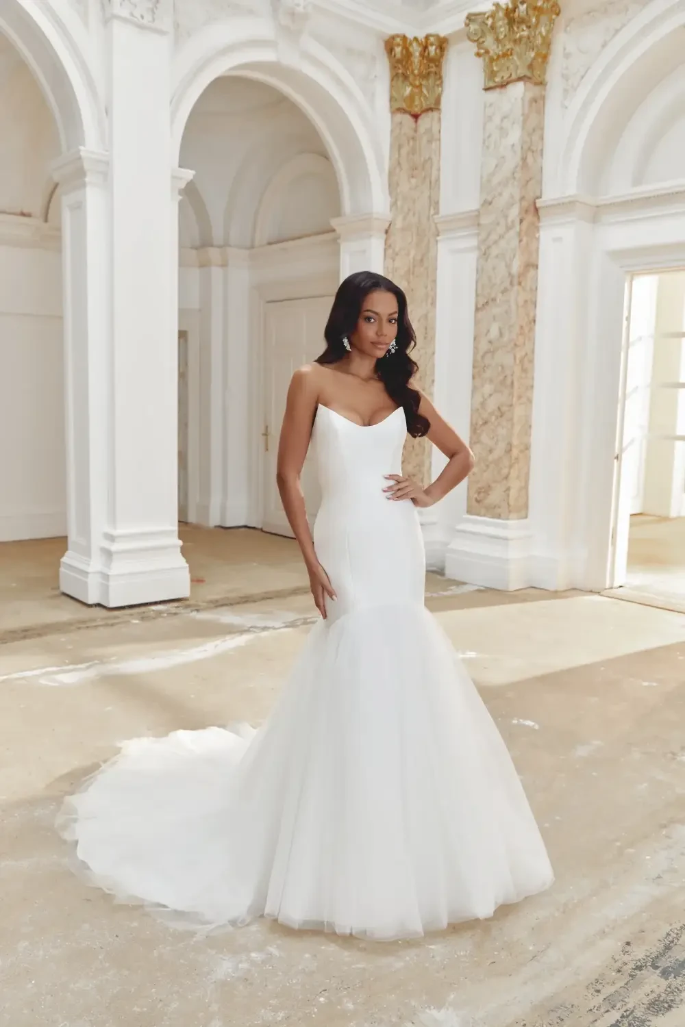 A woman in a fitted white wedding dress standing in a grand, ornate room with white walls and decorative columns.