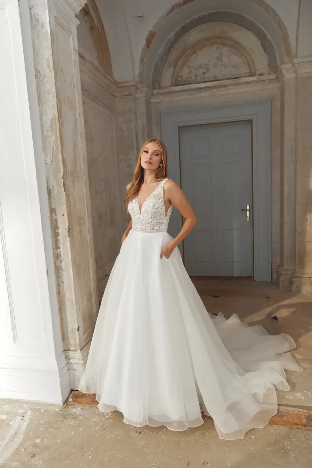 A woman wearing a wedding dress with a deep V-neckline and beaded details, standing in an elegantly lit room with light-colored walls and a closed door in the background. The dress has a full skirt with pockets and a long train.