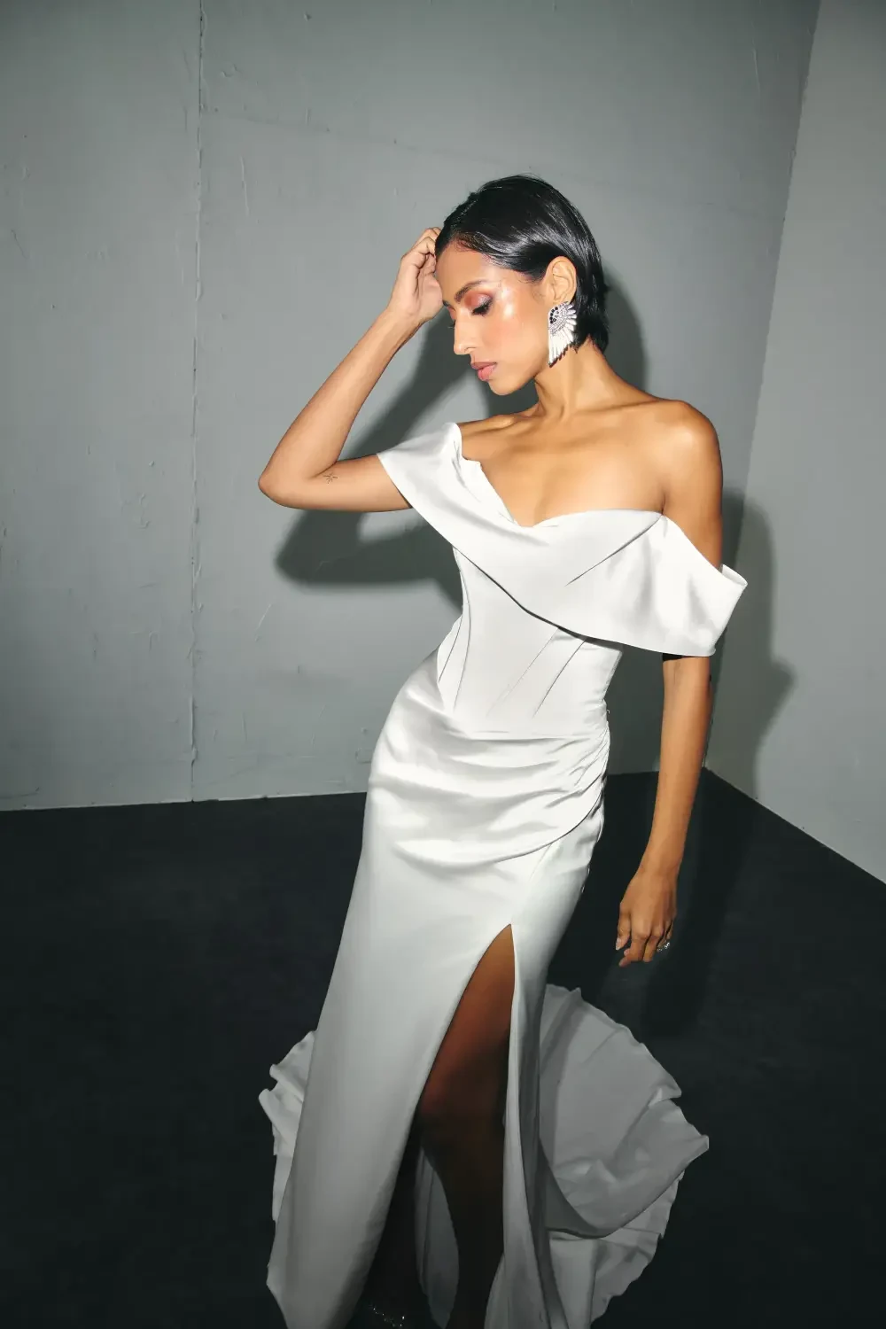 A woman wearing an elegant off-the-shoulder white dress with a high slit, posing gracefully against a minimalist background.