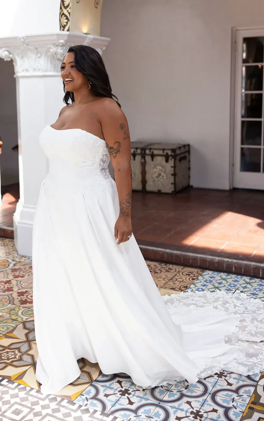 A woman wearing a strapless white bridal gown stands in an elegantly decorated room with patterned tiles.