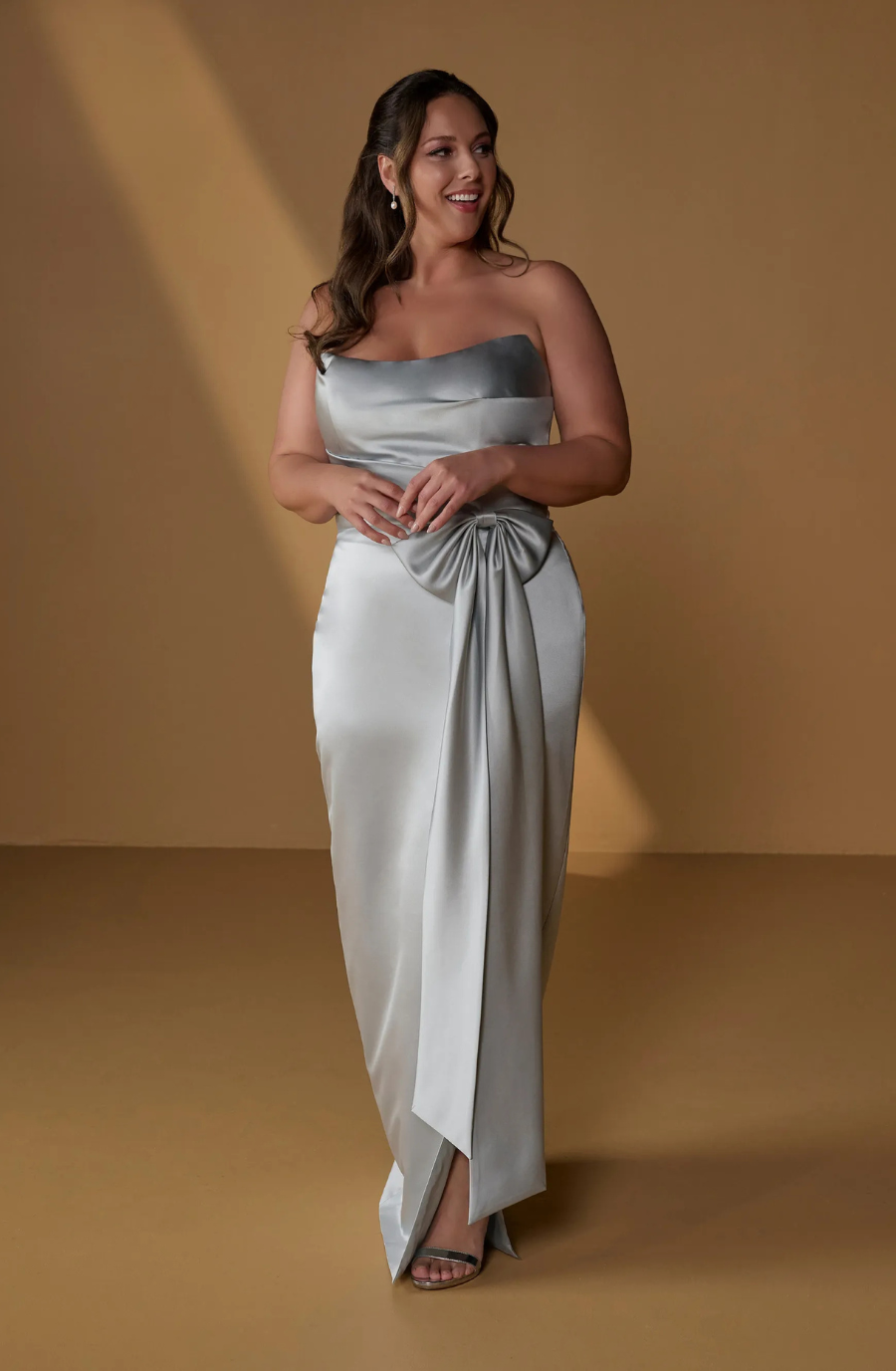 A woman in an elegant silver gown with a bow detail, smiling and standing against a brown background.